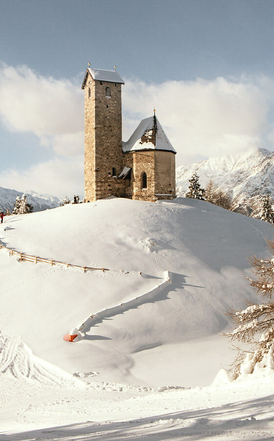 Kirchlein auf Hügel im Winter mit Schnee und Bergpanorama