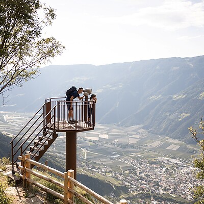 A viewing platform with a telescope on a steep mountainside; it offers a vast panoramic view over the sunny Adige Valley and the surrounding mountain ranges near Merano.