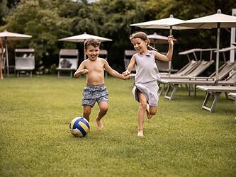 Two smiling children joyfully play football on a well-kept hotel lawn; comfortable sun loungers and umbrellas under trees are visible in the background.