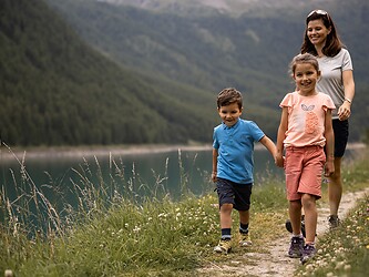 A smiling mother hikes with her two children along a path by an idyllic mountain lake; the children are holding hands, while forested mountains rise into the cloudy sky in the background.