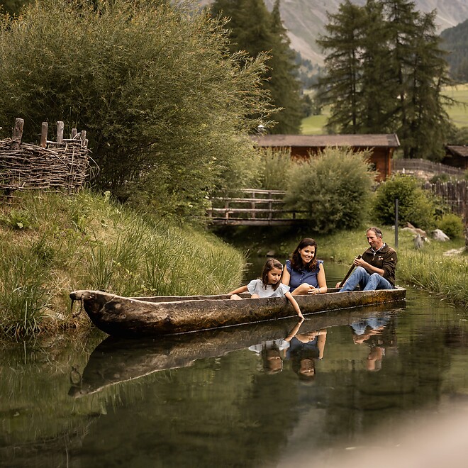 A family with children enjoys a playful boat ride in a rustic dugout canoe on a small pond; surrounded by lush greenery and fir trees, the South Tyrolean mountain peaks rise in the background.