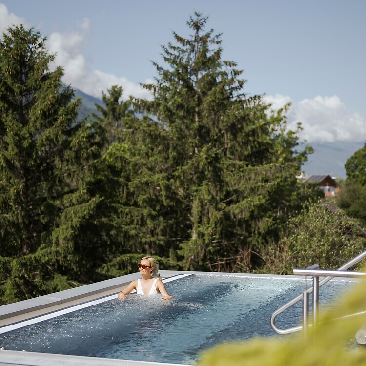 A woman relaxes in a modern outdoor stainless steel whirlpool; the bubbling water glistens in the sun, with tall fir trees and Alpine peaks visible in the background.