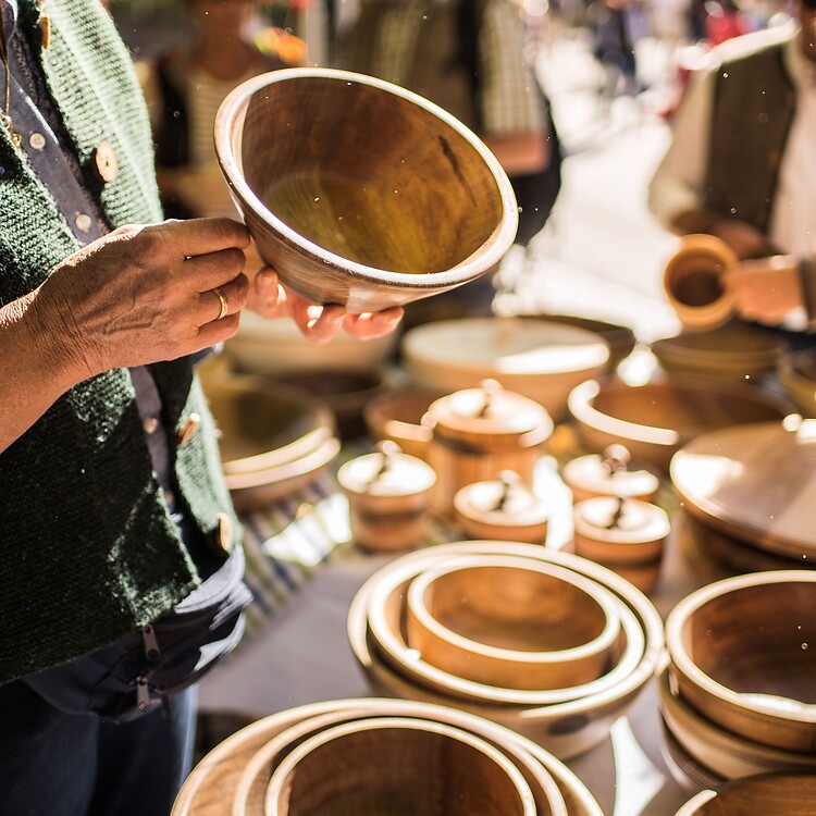 Close-up at a South Tyrolean farmers' market; a customer examines handmade wooden bowls at a market stall, while other visitors stroll through the sunny alleys in the background.