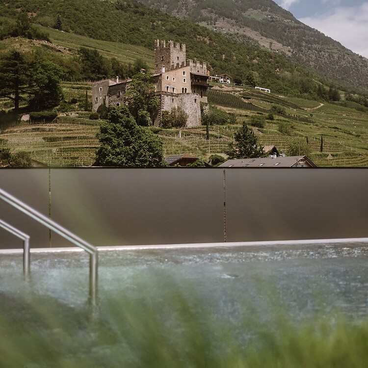 A modern outdoor pool at Hotel Lindenhof in Naturns; in the background, a historic building is visible on a forested mountainside under a blue sky.
