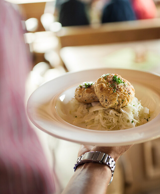 A server presents a plate with two golden-brown pan-fried dumplings on a bed of light sauerkraut, garnished with fresh chives; in the background, the cozy wooden interior of a traditional South Tyrolean tavern is visible.