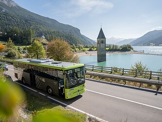 A modern green and black public bus drives along the shore road of Lake Reschen; in the background, the famous historic steeple of Old Graun emerges from the turquoise water.
