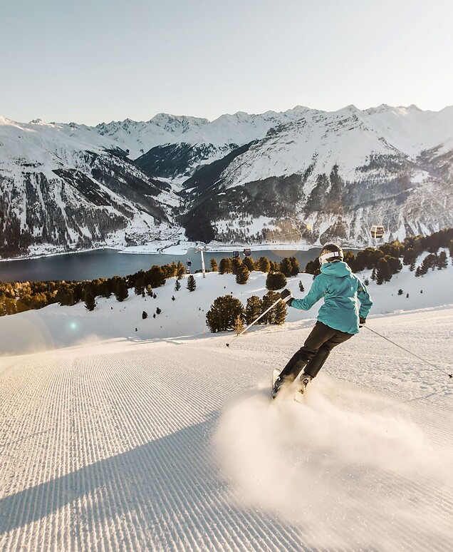 A skier carves down a freshly groomed slope; in the background, a wide panoramic view of Lake Reschen and the snow-capped peaks of the Alps unfolds under bright sunshine.