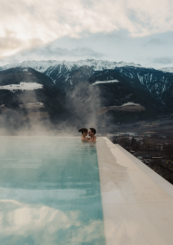 A couple relaxes in a steaming infinity pool; in the background, an impressive snow-covered mountain range stretches out under a cloudy winter sky.