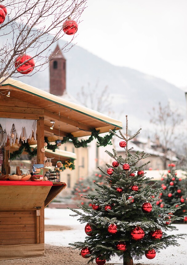 A small Christmas tree decorated with red baubles stands in the snow in front of a rustic market stall at the Sterntaler Christmas Market in Lana.