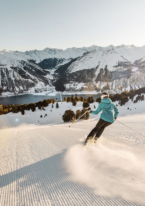 A skier carves down a freshly groomed slope; in the background, a wide panoramic view of Lake Reschen and the snow-capped peaks of the Alps unfolds under bright sunshine.