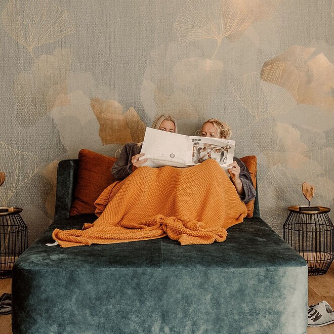 Ein stilvoller Ruheraum im Hotel Jagdhof mit komfortablen, blau gepolsterten Liegen und orangefarbenen Akzenten; die Wandgestaltung zeigt ein traumhaftes Wolkenmotiv in sanften Gold- und Blautönen.