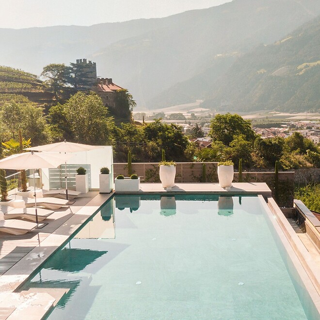 Ein langer, moderner Sportpool im Hotel Preidlhof mit bläulich schimmerndem Wasser, umgeben von einer eleganten Holzterrasse und einer Glasreling, die den Blick auf die mediterrane Vegetation freigibt.
