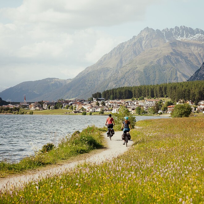 Zwei Frauen biken am Reschenseeweg vorbei an blühenden Wiesen
