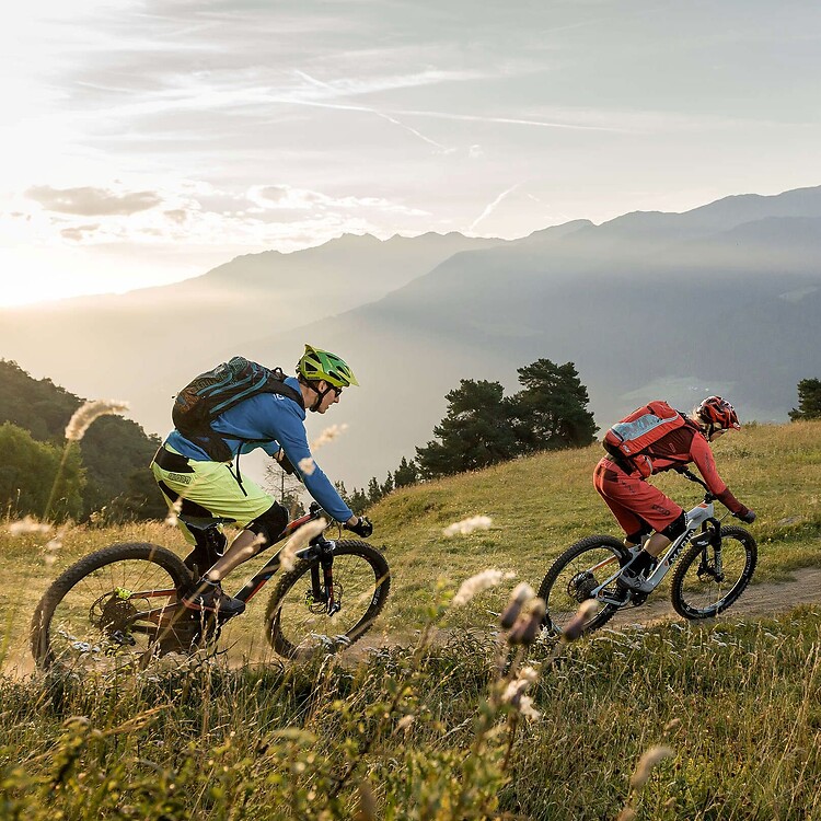Zwei Mountainbiker fahren während der Goldenen Stunde über eine Bergwiese, im Hintergrund erstreckt sich eine weite Gebirgskette.