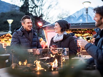 Three smiling friends enjoy the wintry atmosphere at the Merano Christmas Market, warming themselves together around an open outdoor fire pit.