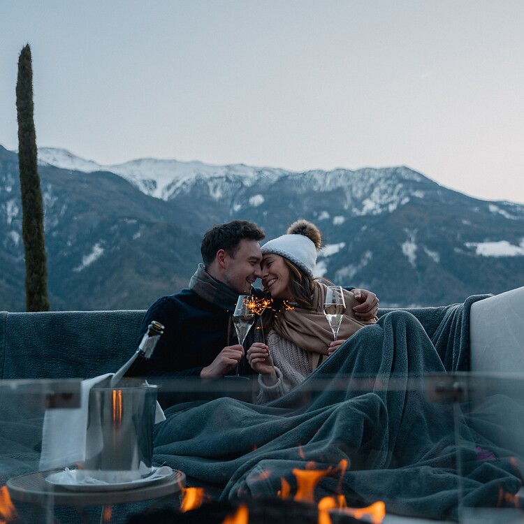 A smiling couple toasts with wine glasses at winter twilight; they are sitting cozily by an outdoor fire pit with a view of the snow-capped mountain peaks.