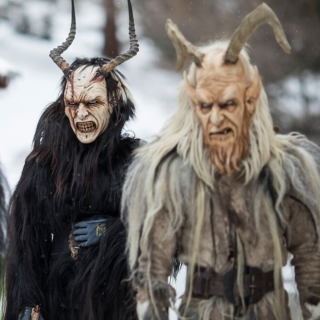 Two horned Krampus figures with long hair and terrifying faces stand in a snowy winter landscape.