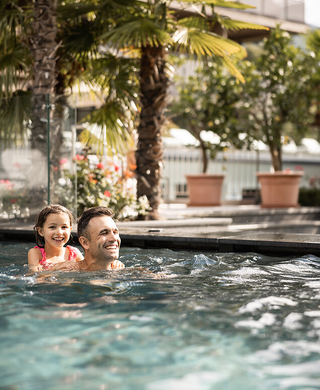 Ein lächelnder Vater schwimmt mit seiner kleinen Tochter auf den Schultern in einem Außenpool, umgeben von Palmen und mediterranen Pflanzen im Gartenbereich eines Hotels.